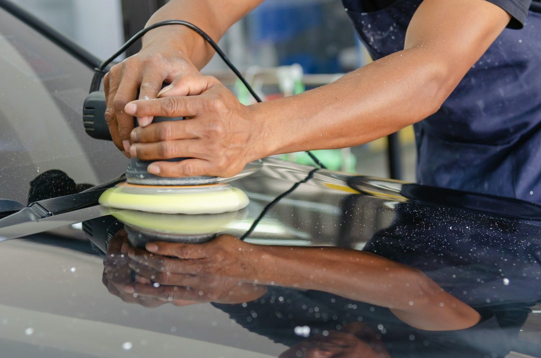 Detailed view of a worker's hands polishing a car's hood using a buffer in an auto workshop.